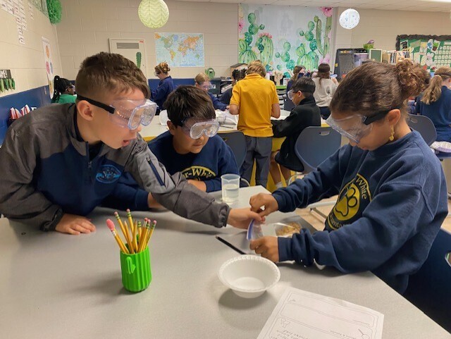 Kids wear goggles. They work on a table with pencils, a bowl, and paper. A map is on the wall.