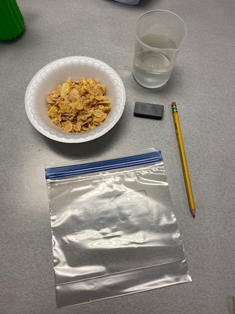 A table has a bowl of cereal, a clear plastic bag, a yellow pencil, a small gray eraser, and a glass of water.