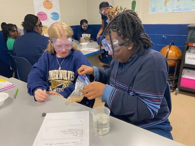 Two students with goggles on conduct an experiment in a classroom. One pours liquid into a cup.