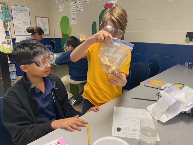Two children wearing safety goggles examine a clear plastic bag filled with water, seated at a table in a classroom.