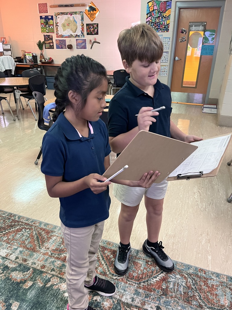 Two students in blue shirts hold papers and a clipboard in a classroom with chairs and tables.