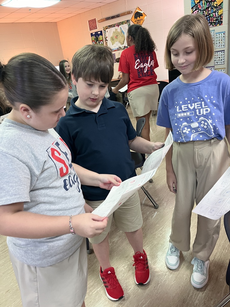 Three children stand together, each holding a piece of paper. One wears a navy polo, another a gray shirt, and the third a blue top.
