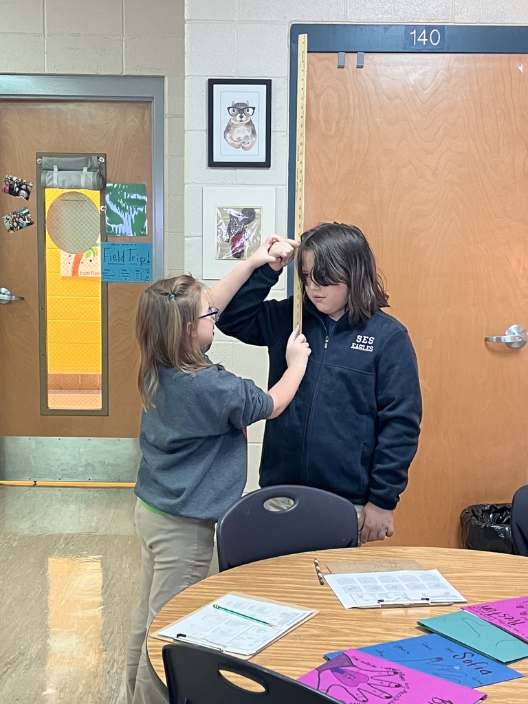 Two students measure a pole next to a table with papers and chairs in a room with wooden doors.