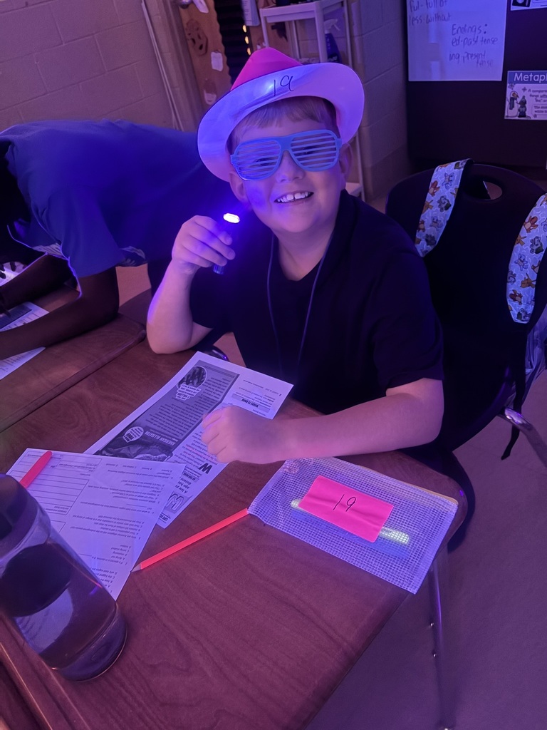 Child at desk with flashlight, smiling, wearing a hat and glasses. Papers, pens, and a plastic container on desk.