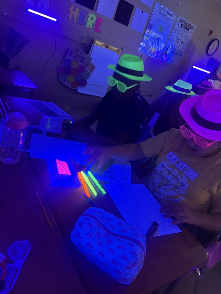 A person with a green hat sits at a desk with others, all wearing hats. They are using glow sticks, and the room is dimly lit.