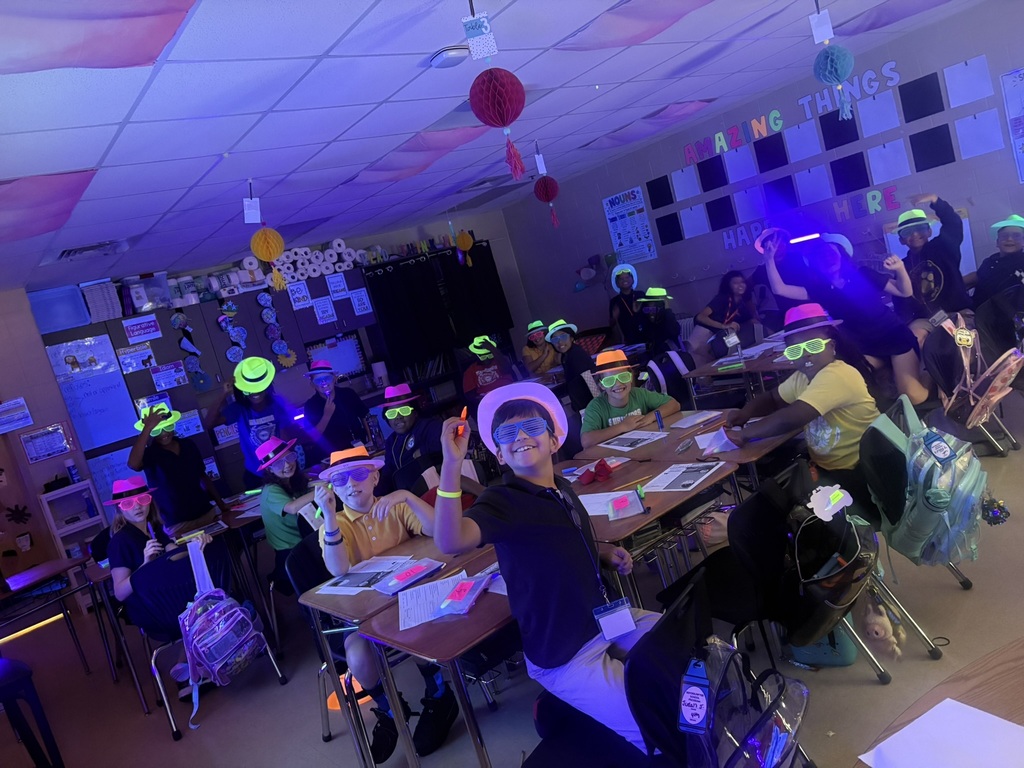 A classroom with students seated at tables wearing light-up hats. Purple lights illuminate the room.