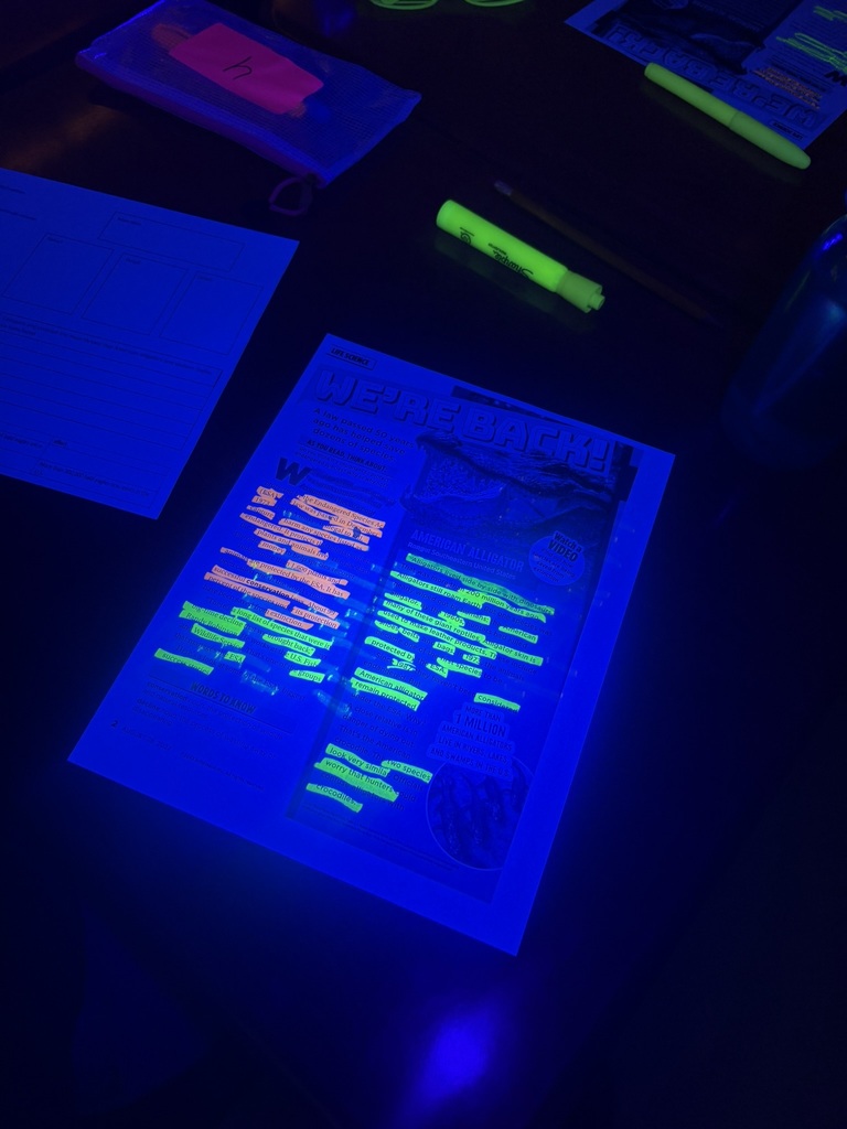 Dark room with a table containing papers and sticky notes. A paper with highlighted text is under a UV light.