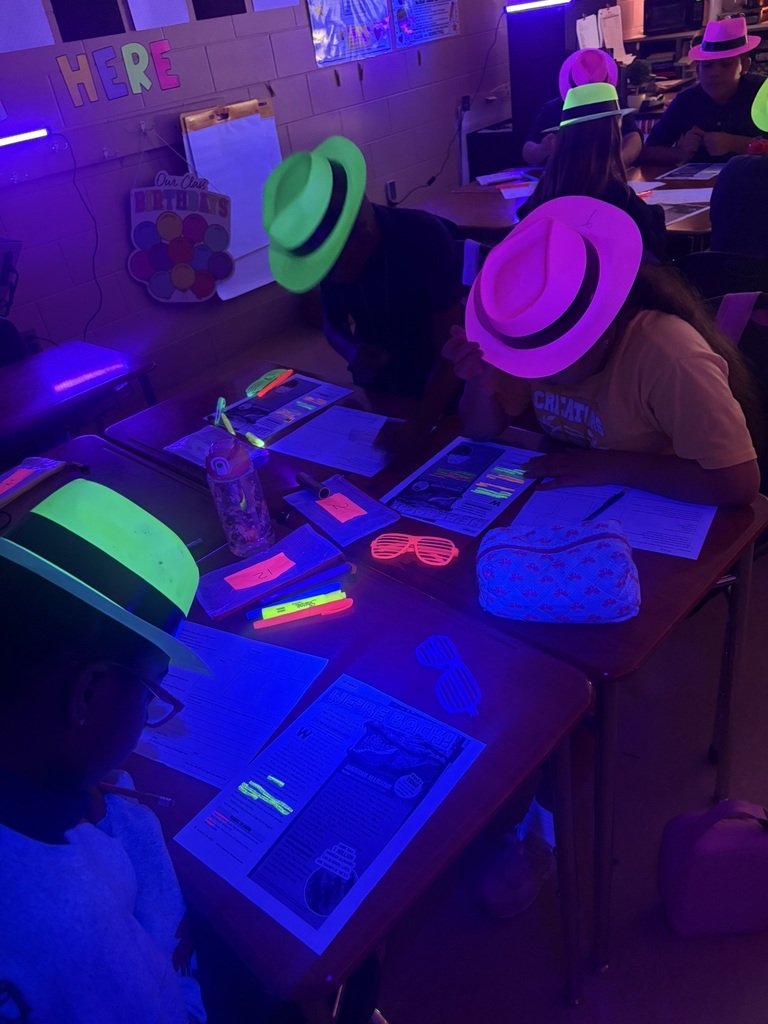 Students seated at tables wearing neon green hats and pink hats, with papers and pens. Fluorescent lights and an illuminated wall.