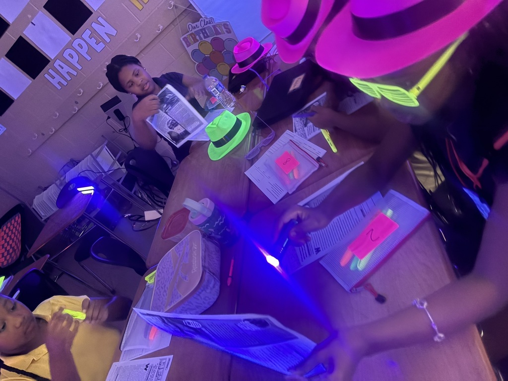 A group of people at a table wearing glow-in-the-dark accessories and reading papers under purple lights.