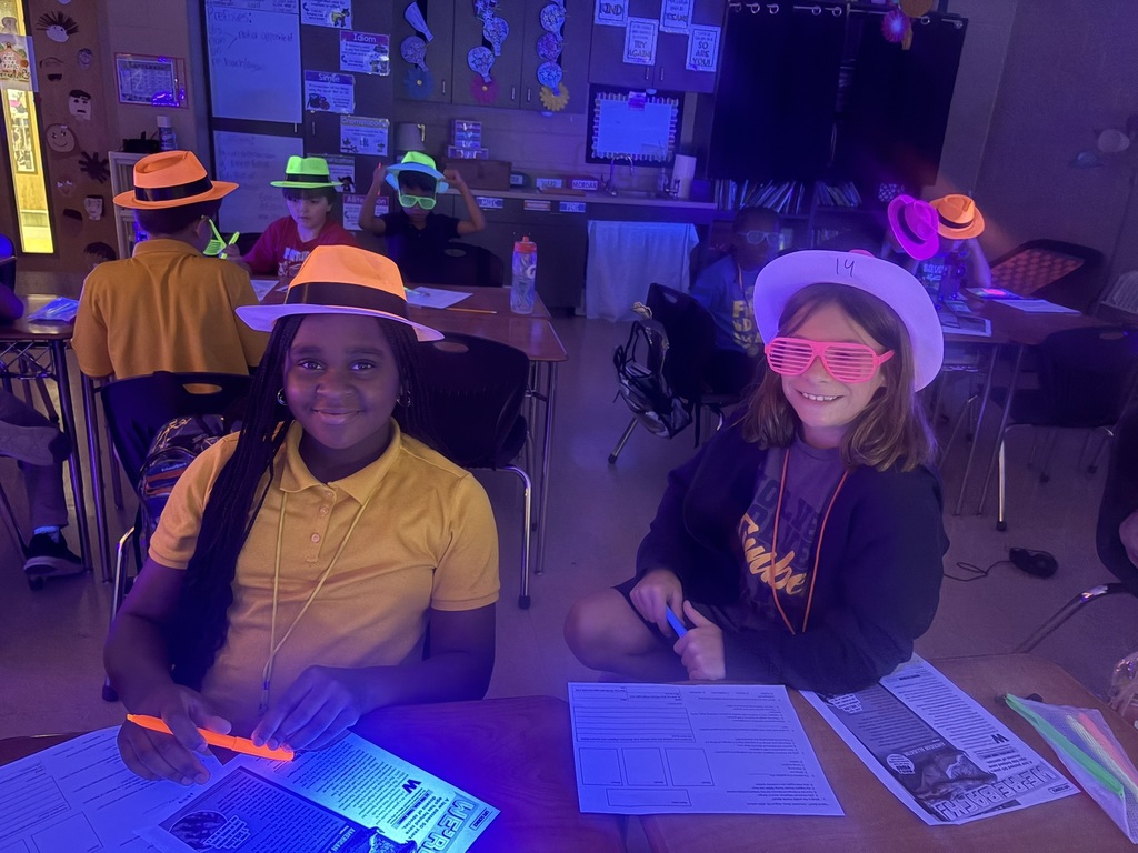Two girls in hats and glasses sit at a desk, surrounded by other students. One holds a pen.