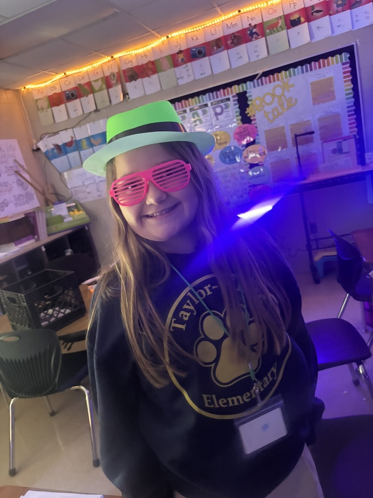 A young girl in a green hat, pink sunglasses, and a Taylor Elementary sweatshirt smiles in a classroom.