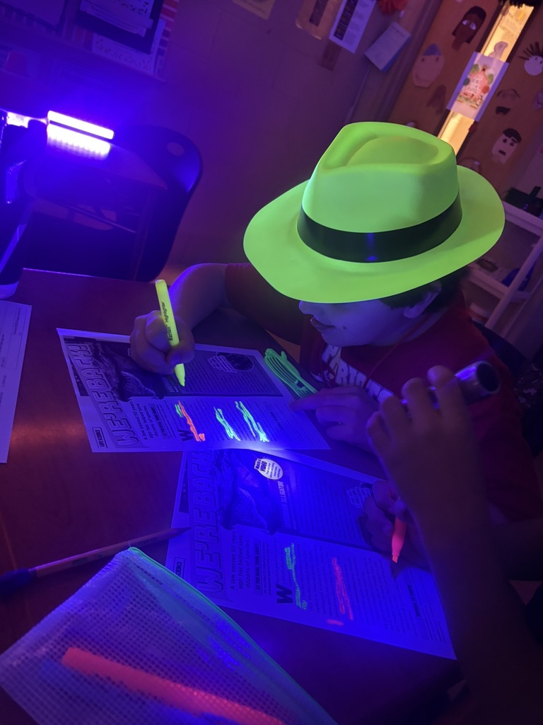 A person with a green hat uses a neon green pen on a worksheet. The room is dimly lit with purple light.
