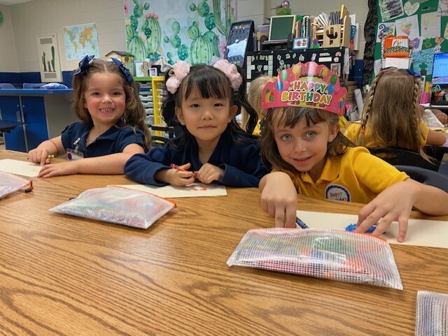 Three children sit at a table, one with a party hat. A girl holds a pen and paper.