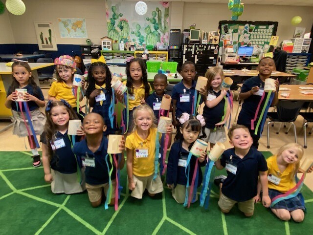 A classroom filled with children, some standing and some sitting, holding craft items and posing for a picture.