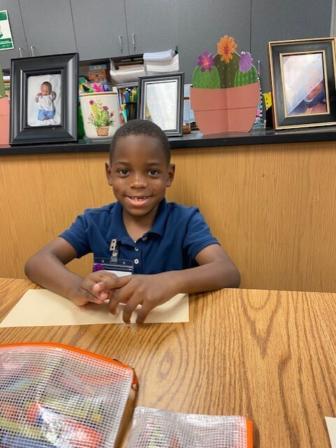 A child sits at a desk with a smile, hands resting on paper. Behind, a wooden counter has picture frames and potted plants.