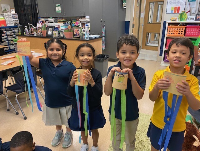 Five children in matching uniforms hold colorful paper creations in a classroom. They smile and stand together.