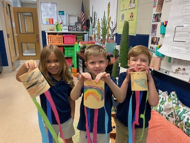 Three children hold up handmade kites, smiling for the camera. Behind them, shelves hold colorful items.