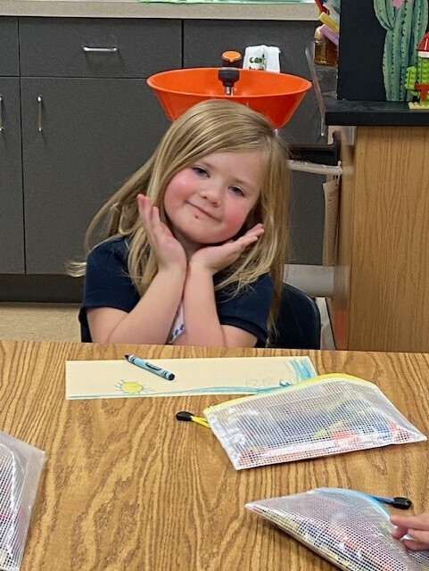 A young girl with blonde hair sits at a desk, smiling, with a colored pencil in her hand.