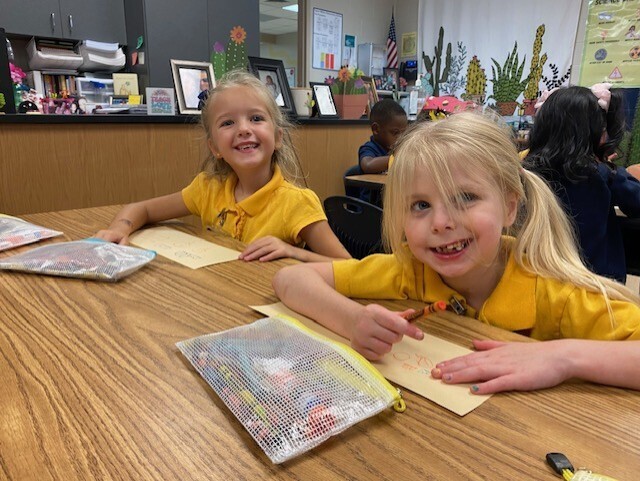 Two children, one with a ponytail, sit at a desk, smiling. Behind them, a room with desks, a bulletin board, and potted plants.