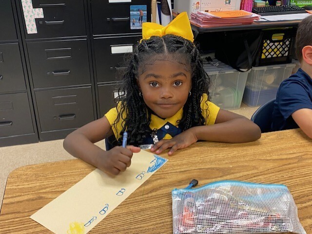 A young girl with a yellow bow sits at a desk, holding a pen and writing on paper.