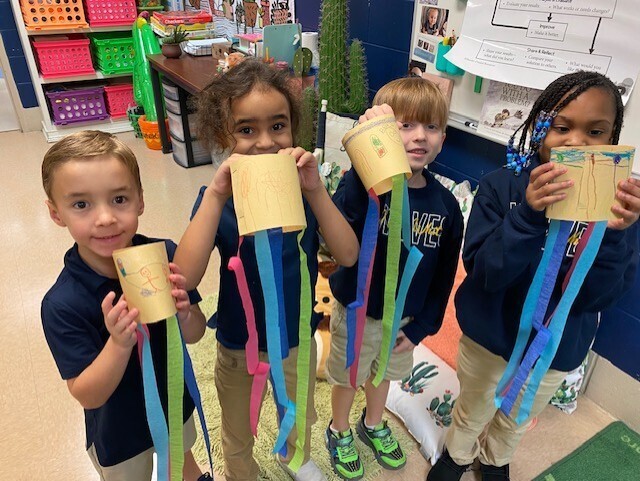 Four children, in navy blue uniforms, hold paper cups with multicolored ribbons. They stand in a classroom.