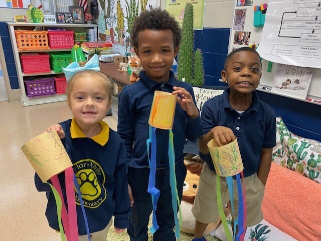 Three children in a classroom hold colorful paper crafts. Background includes shelves, a cactus, and a whiteboard.
