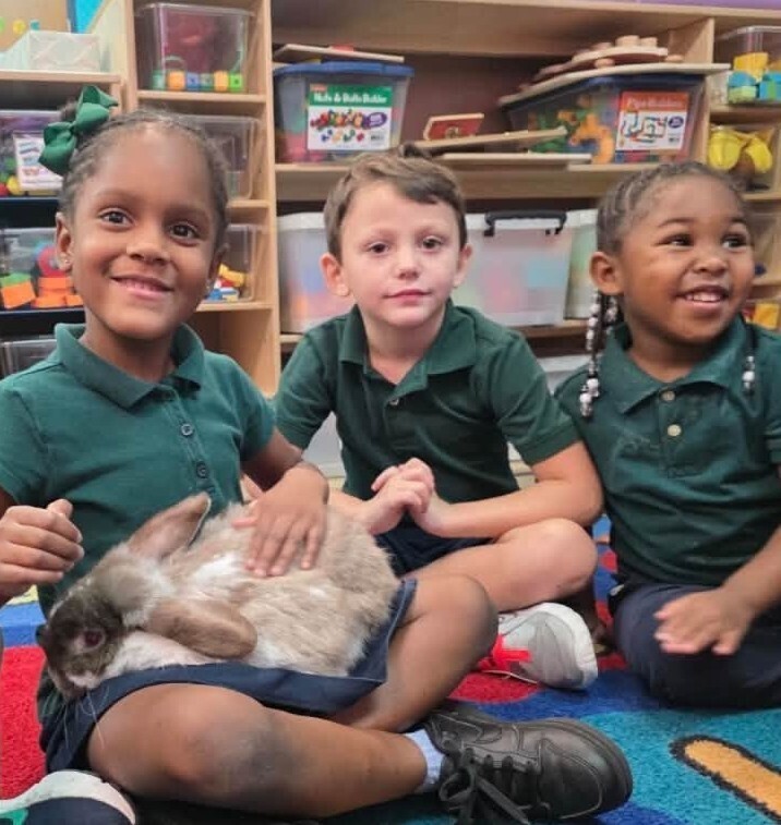 Three Pre-K students sitting on the floor, with one holding a bunny in her lap