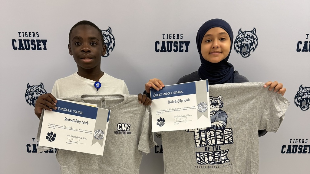 Two children in a school setting hold certificates and gray T-shirts with "Tigers" printed on them.