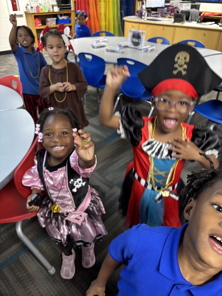 Four children in pirate costumes stand in a classroom. Three of them have raised hands, and one wears a pirate hat.