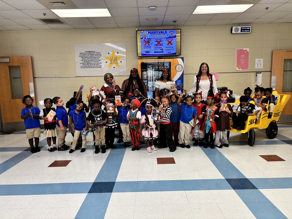 A group of children dressed in costumes stands in a corridor with adults. A yellow toy cart is nearby.