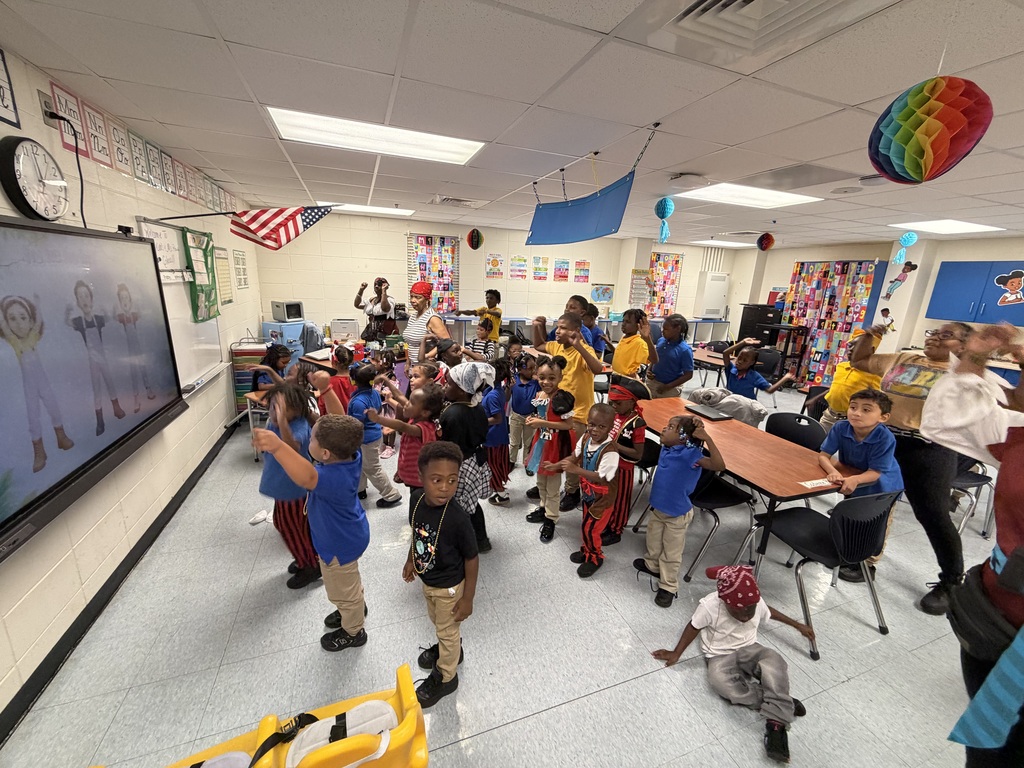 Kids and adults are in a classroom. They are gathered around a large screen and some are standing or sitting.