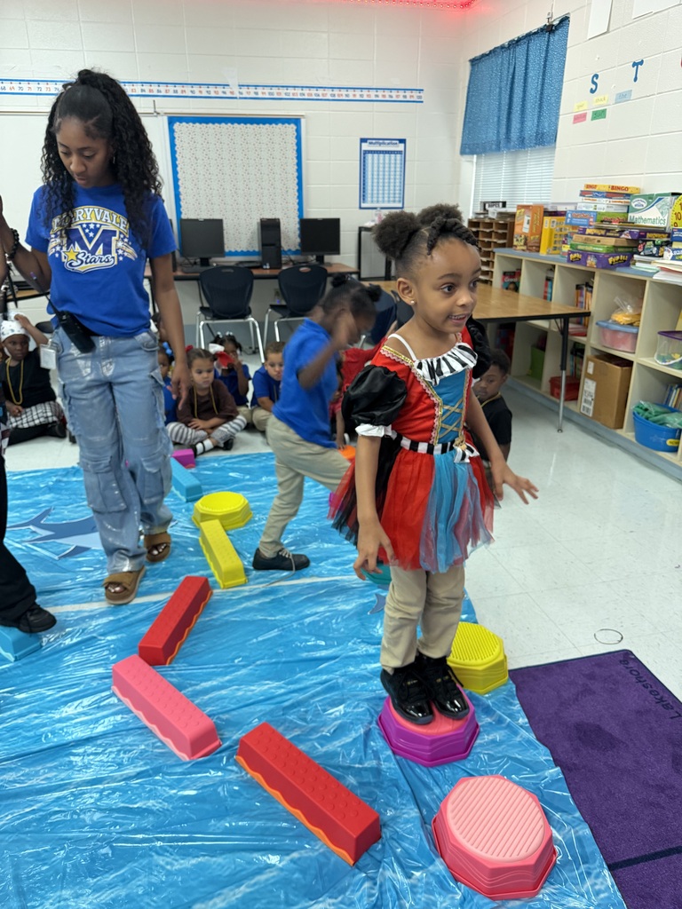 Kids in a classroom standing on various colored foam blocks on a blue mat. A girl in a red and black outfit is smiling.