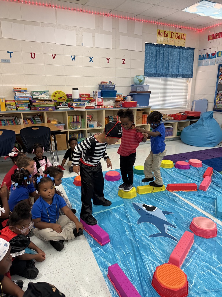 Children play on a floor covered with colorful mats, with some standing on blocks. Others sit nearby.