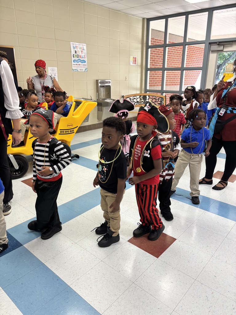 Children in a school hallway, several in pirate costumes, with adults nearby and a yellow vehicle in the background.