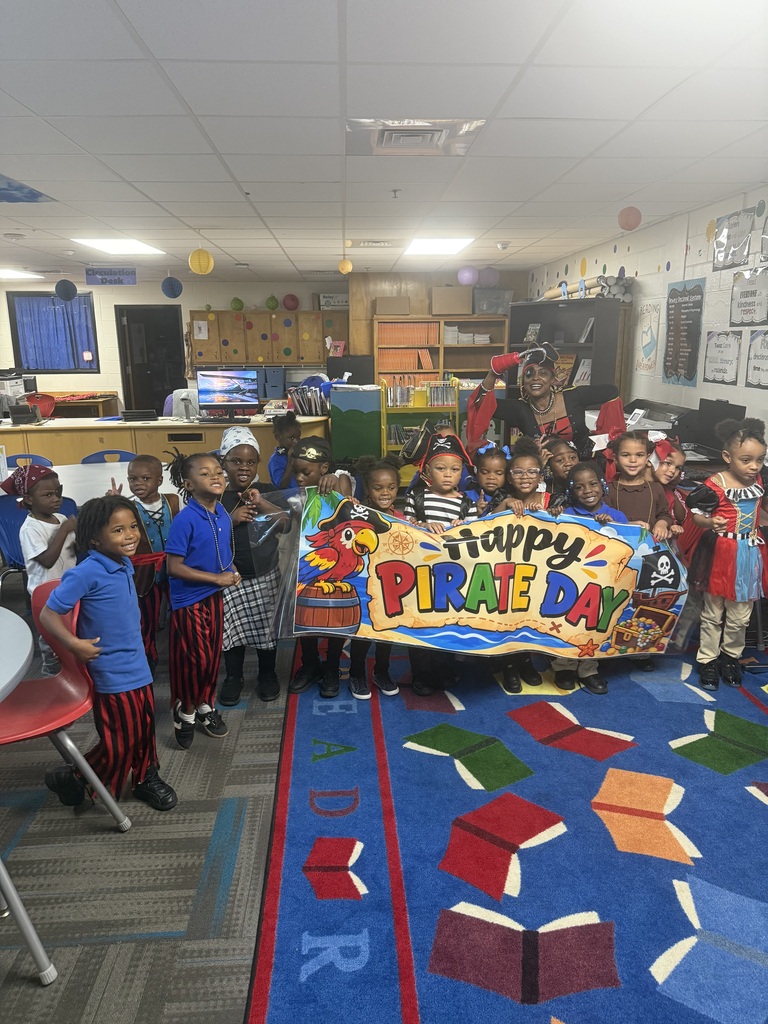 A group of children stands in a classroom, holding a banner that says "Happy Pirate Day". Colorful books are on the floor.
