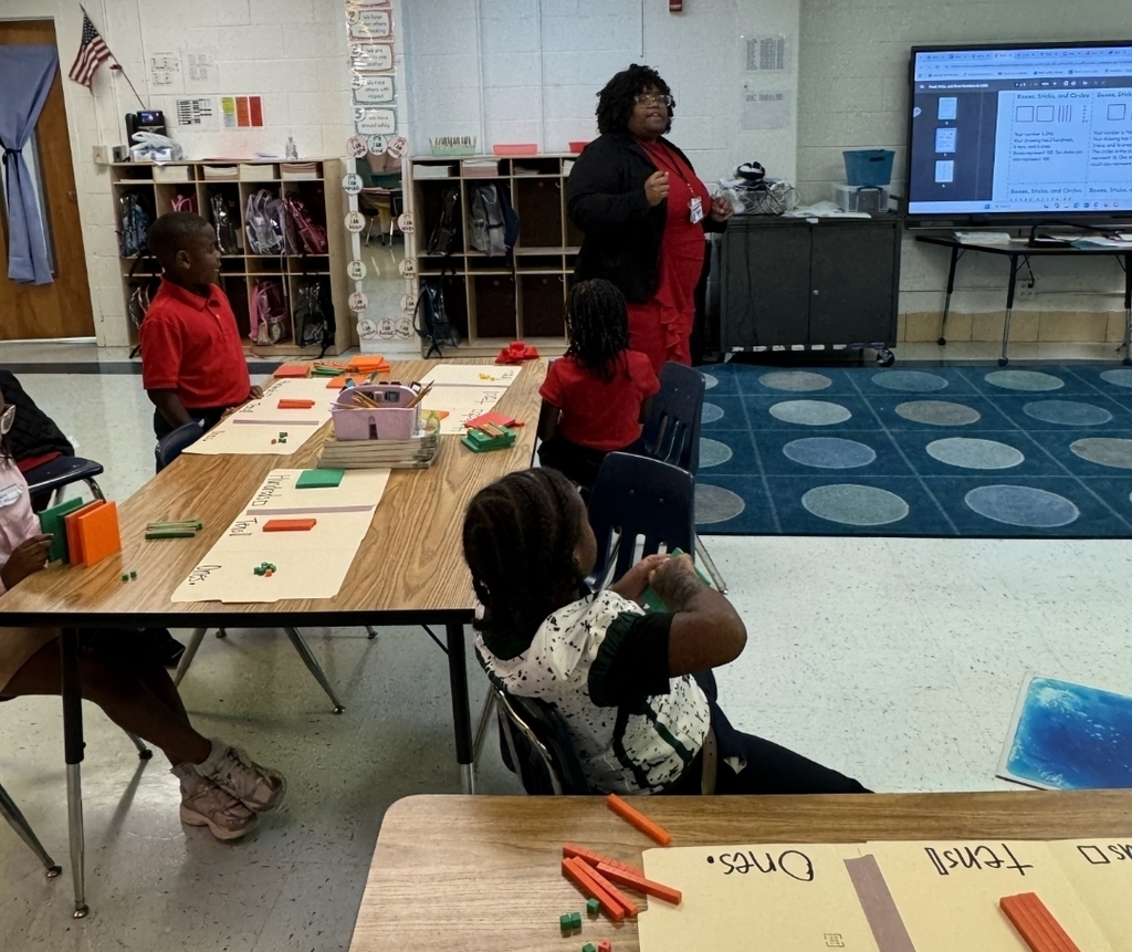 A classroom with students seated at desks. A teacher stands at a table, explaining something with a computer screen behind.