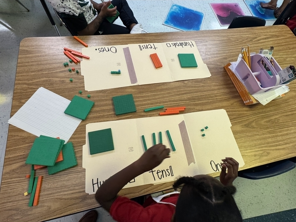 A child seated at a table with educational materials, including green shapes, pencils, and paper, working on a math activity.