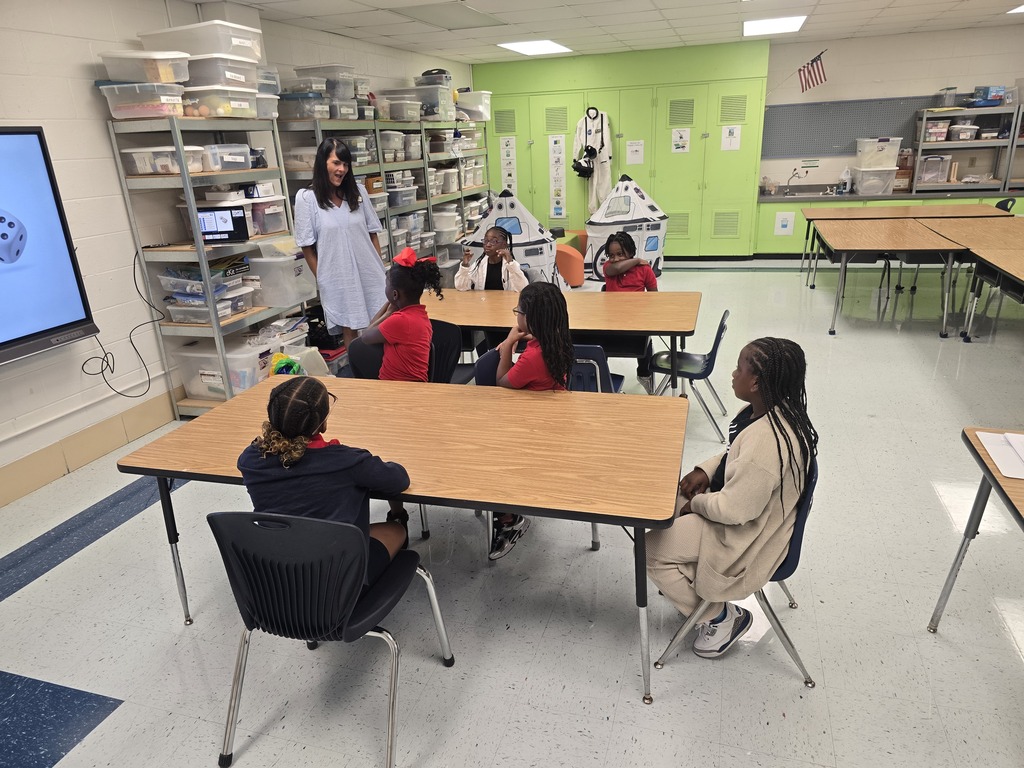 A woman stands before a group of students seated at tables in a classroom. Shelves and cabinets are in the background.