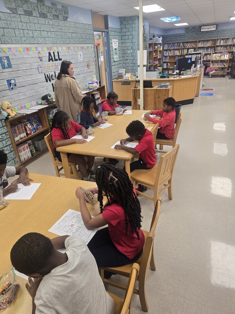 A classroom with several children seated around tables, working on papers. A woman stands near a bookshelf in the background.