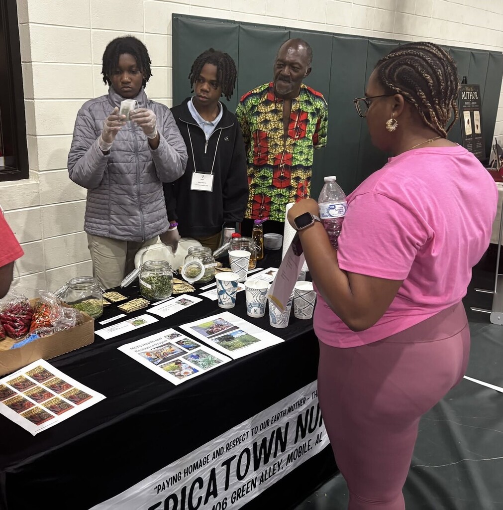 A man and two students working behind a booth full of tea and peppers, with a customer looking on