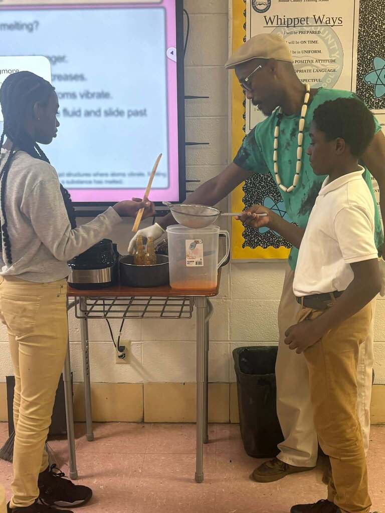 Two students straining peppers into hot sauce as a man watches