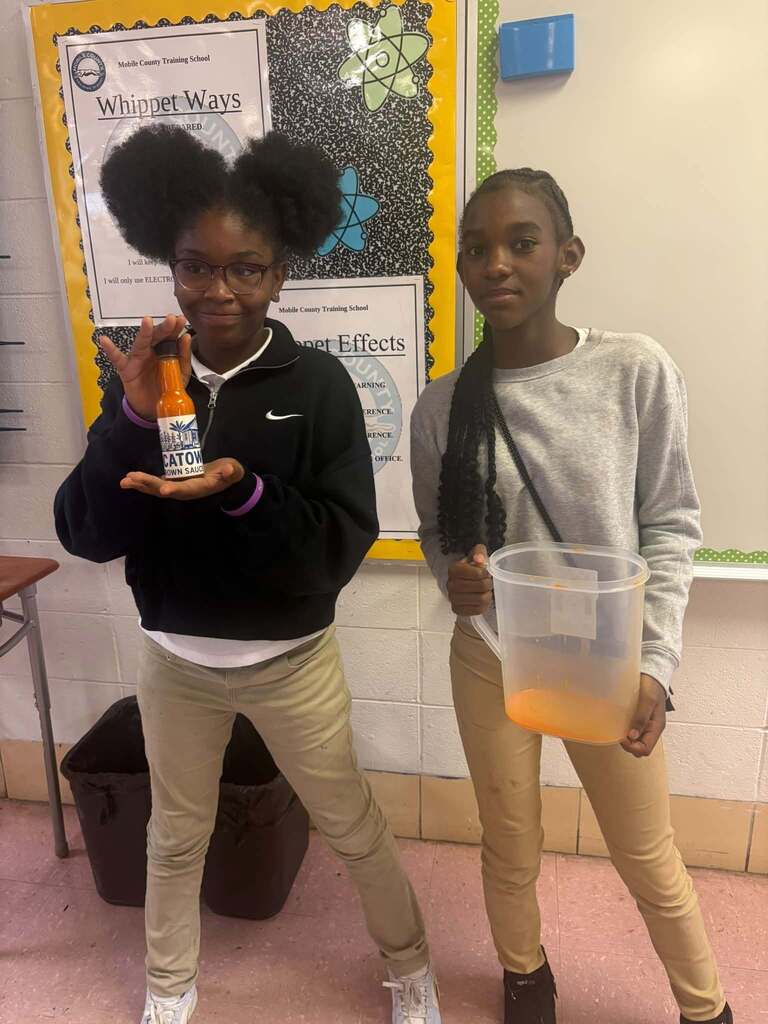 A girl holding a jar of hot sauce and another holding a pitcher