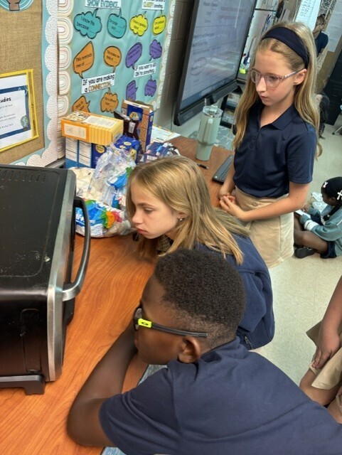 Four students in a classroom setting, one leaning over a table with a microwave, another seated, and two standing nearby.