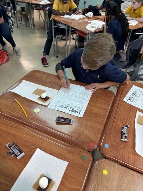 In a classroom, a student writes on paper at a desk with a pen, pencils, and other items.