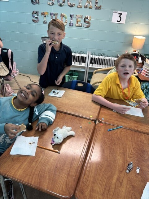 Students sit at desks with one using a toothbrush, another with a toy, and a third holding paper.