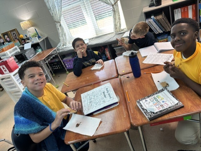 Four children are seated at desks in a classroom, with papers, notebooks, and pens in front of them.