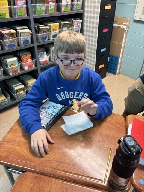 A boy with glasses sits at a desk eating a cookie. A book and napkin are on the desk.