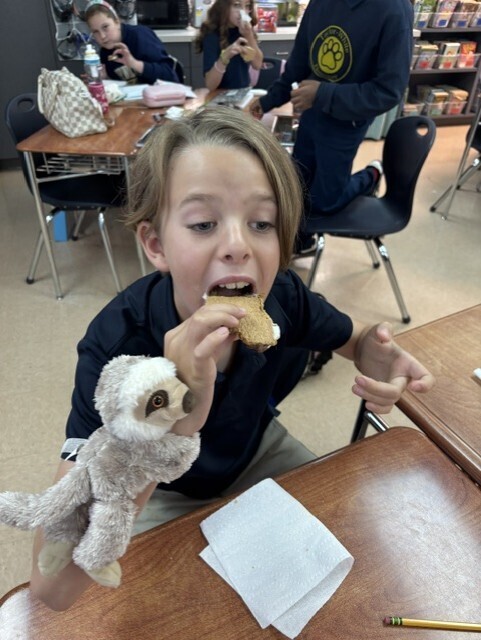 A child in a blue shirt eats a sandwich with a stuffed animal in a classroom.