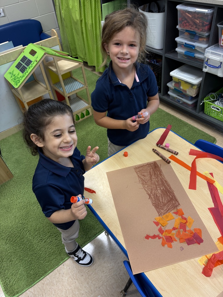 Two girls in matching uniforms work on a paper art project at a table in a classroom.