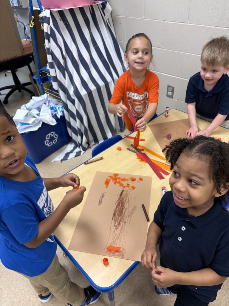 Four children seated around a table, engaged in an art activity, with one child holding a brown paper.